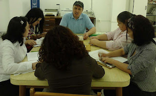 Foto dos alunos sentados em volta de uma mesa. Na ponta da mesa está o professor Airton vestindo uma camisa gola pólo azul e boné preto.a sua direita está Marmis lendo a apostila de braille, logo em seguida está a Cida, de camisa branca, conversando com o professor Airton.  Na outra ponta da mesa está a Chica, vestindo camisa marrom, cabelos soltos. A direita da Chica está a professora Izabel, de camisa listada, faixa nos cabelos, escrevendo na reglete. Em seguida está a Kelli de cabelo amarrado num rabo de cavalo, vestindo camisa com listas branca e rosa. 