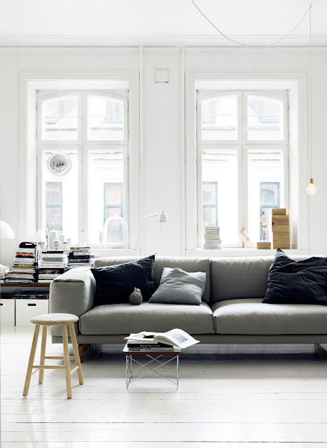living room with a gray sofa with gray and black acecnt pillows, a side table full of books, a stool being use as a tiny coffee table and tall windows