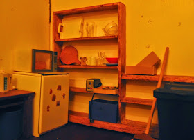 Modern miniature scene of a holiday house kitchen at night, with a bar fridge, microwave and shelving holding various items of crockery and kitchenware.