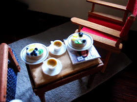 Aerial view of a miniature holiday house scene showing a sitting area with a 1950s-style lounge chair and directors chair . Between them is a piano stool set with two flans on plates and two cups of tea on top.