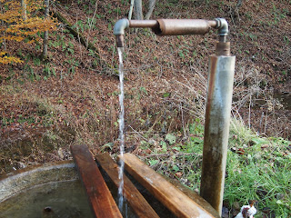 A pipe runs up from ground, then turns, so water flows into basin, splashing on wooden boards as it does.