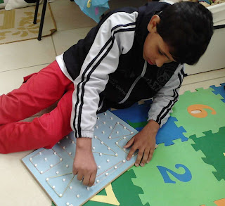 Foto do Eduardo sentado no tapete dos números e brincando de formar figuras no geoplano