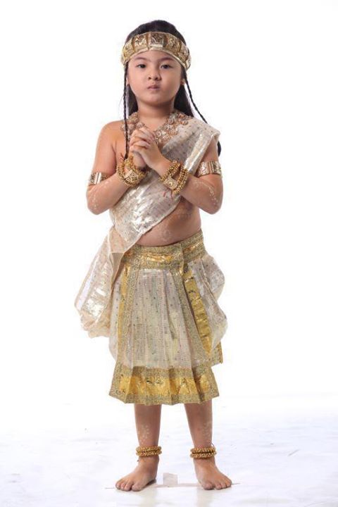  A child dressed in white precolonial attire, adorned with gold necklaces, headbands, armbands, legbands. Hiis arm tattoos are glowing in gold.