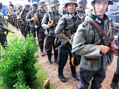 Line of 1/6 scale German soldiers marching in a diorama of an army post on display at a scale model exhibition.