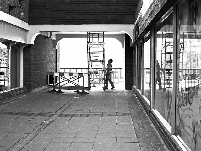 Man moving a small scaffold tower while working on repairs in shopping centre. Black and white with reflections in windows.