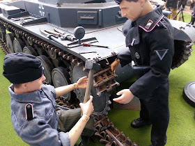 Two 1/6 scale German soldiers fixing a tank tread in a diorama of an army post on display at a scale model exhibition.