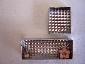 Two silver-coloured embossed metal dolls house wall boxes, one rectangular and one square, with a perfume bottle and frangipani in the lower one.