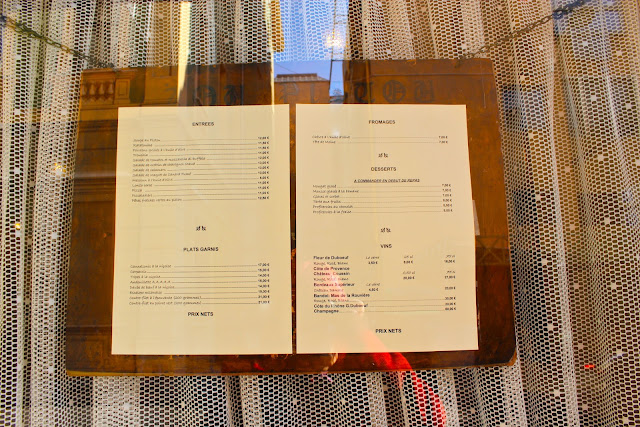 Menu chained up against a lace curtained restaurant window. A regional speciality is 'Socca', chickpea flour crepes.