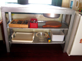 Modern miniature metal kitchen bench unit displaying a selection of kitchenware underneath.