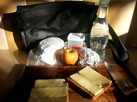 Cheese on toast on a chopping board. Behind it on the kitchen bench are various tinfoil packages and plastic containers, an apple, and a bottle of water.
