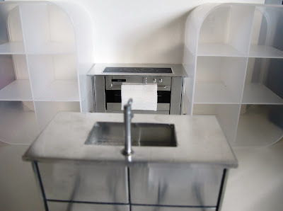 Modern dolls' house miniature set up with a metal stove and kitchen unit with sink, and two white perspex shelving unit.