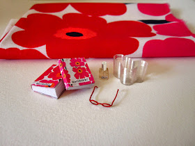 Miniature Marimekko books, red reading glasses, Dior perfume bottle and  Aalto vase arranged in front of a piece of full-sized Unikko fabric.