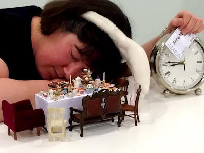 Woman wearing rabbit ears, asleep in front of a dolls' house miniature table set for tea, holding a large clock.