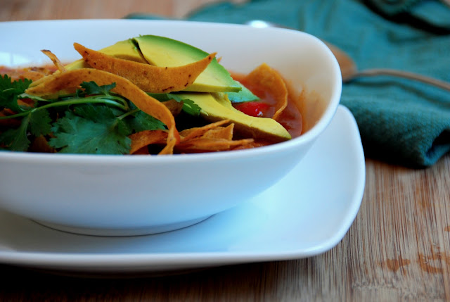 tortilla soup topped with sliced avocado, tortilla strips, and cilantro in white bowl on white plate in front of a spoon on a blue napkin