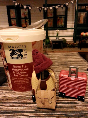 Two modern dolls' house miniature suitcases next to a container of ice cream, on a dining table in from of a dolls' house miniature school.