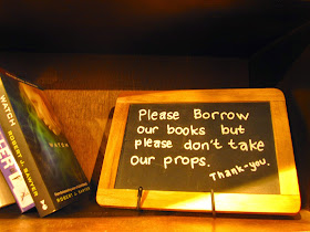 Blackboard sign on a shelf of books at The Little Library in Melbourne Central: 'Please borrow our books, but please don't take our props. Thank you.'