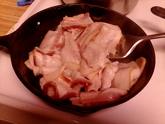 Iron Skillet with Applewood smoked bacon frying with silver spatula in pan.