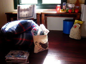 Inside of a miniature holiday house, showing a doona rolled up on the floor next to a pile of magazines and a bag of knitting. On the table behind is a selection of miniature grocery items, and a bag of groceries is on the floor underneath,