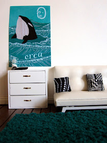 Modern dolls' house lounge room with teal flokati rug, white sofa and drawers, black and white cushions and orca art poster