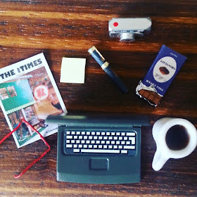 Flat lay of  a laptop,pair of reading glasses, magazine cover, pen and post it notes, digital camera, bar of chocolate and a mug of coffee.