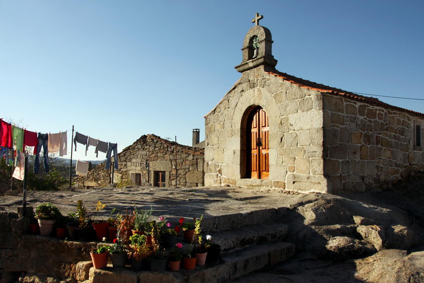 Vista da capela, assente em rocha de granito, com uma eira na frente e roupa a secar. Em primeiro plano vários vasos pequenos nos degraus de acesso ao páteo