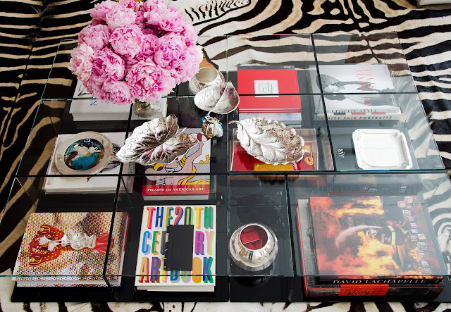 cool coffee table from Cassina that comes in sections letting you decorate the top and bottom. On the bottom there is a collection of books with brightly colored covers and on top there is a flower arrangement made of pink peonies and silver leaf shaped tray. The table is on top of multiple zebra print area rugs. 