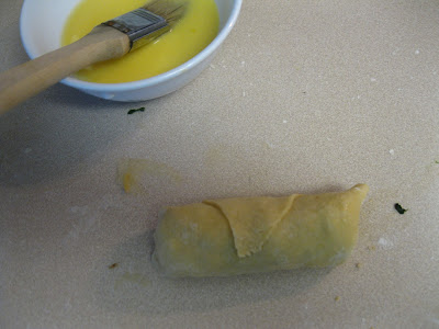 A pastry roll on a countertop next to a small white bowl filled with a beaten yellow mixture, likely egg, with a brush resting on the bowls edge.