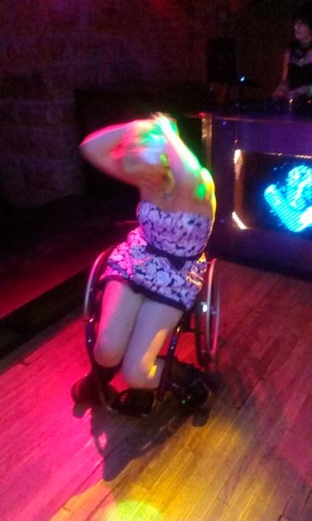 Blonde woman in wheelchair making dance moves, with colored club lights and blurred movement