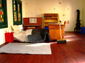 Miniature school house interior showing unmade bed on the floor with coffee mug next to it and a broom propped up at the end of it.