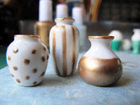 Three white dolls' house vases decorated with gold nail varnish: one spotted, one striped and one dipped, each with gold rims.