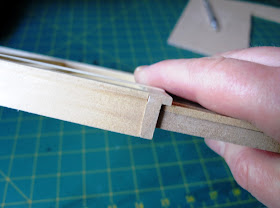 Hand holding up a piece of dolls' house kit wall, with the edge to the camera showing how there is space to add siding  and have room against the door frame.