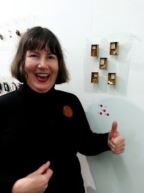 Woman looking happy and making a thumbs up sign in front of five brooches with four red dots. SHe is pointing at a red dot brooch on her jumper.