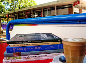 Outside cafe table containing a pile of six new books and a cup of coffee, overlooking Dickson Post Office.