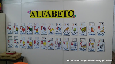 Imagem mostrando o quebra-cabeça do alfabeto colado na parede da sala 2. Sobre cada letra do alfabeto, foi colocado também a letra correspondente em braile e Libras. As palavras referentes as letras tem velcro na parte de trás para ficarem fixas na parede.
