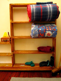 Miniature shelving with a rolled up doona on the top shelf and a folded vintage quilt on the shelf below, On the bottom shelf is a pair of green socks and a pair of gumboots.