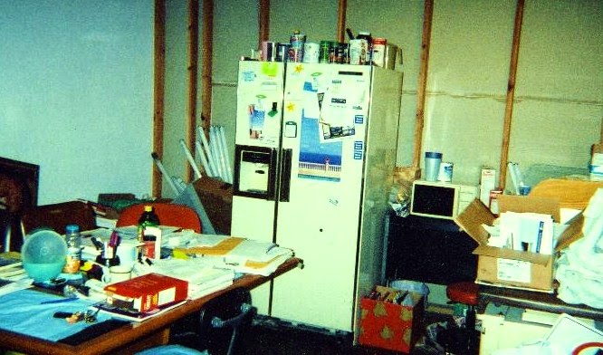 A room with a table and a large refrigerator. It is extremely cluttered, with papers, books, bottles, boxes and so on every surface and stacked in corners