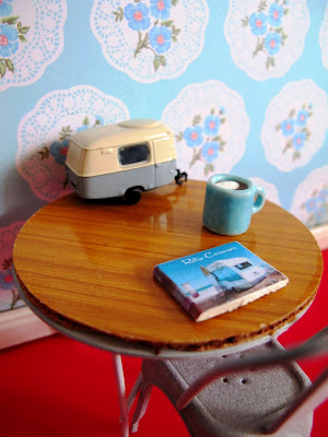 Modern dolls' house miniature scene of a retro table and chair. On the table is a model caravan, a mug of coffee and a book on retro caravans.