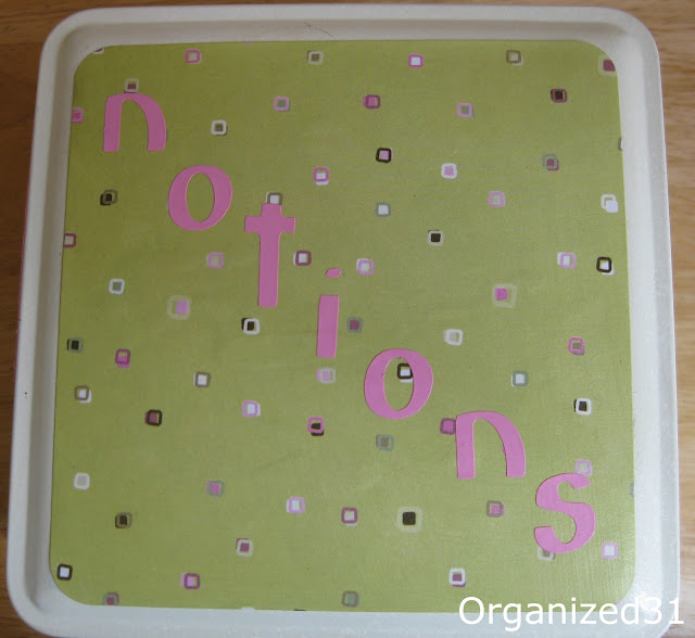 top view of tin decorated with green paper with white, brown and pink squares and pink stripe at tope and bottom and pink letters spelling out "notions"