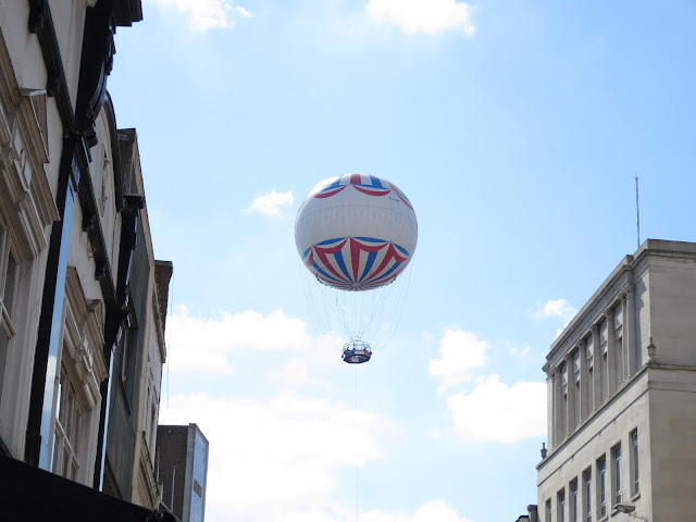 White passenger balloon with red and white patterns, basket below, abpve city street.