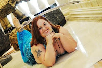 Author laying onfloor with a slim scimitar balanced on her head. Hair is auburn and drapes over her shoulders, baby dragon tattoo on arm, hands folded under chin with legs crossed in the background. Costume is turquoise with bedlah bra, belt, and chiffon pants