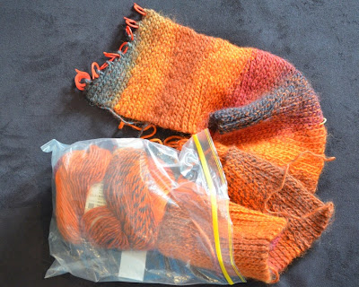 An unfinished scarf in warm reds, oranges, and charcoal looks like it is trying to shuffle out of a plastic snap-lock bag.  The working row is covered wihth red stitch markers.