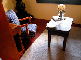 Dolls house miniature sitting area with a 1950s-style reading chair and a small shabby coffee table with a vase of daisies on it.