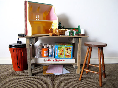 Modern doll's house miniature scene of a workbench with a dolls' house on top, surrounded by tools, paint and miniature furniture.