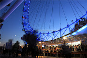 People line up to get on board, while London Eye appears to overpower the . (london eye lit in its blue light overpowers the moon while people line up to get on board)