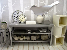 View of a modern dolls' house miniature homeware shop in grey and white. On display is a stack of cafe stools and a grey metal shelving unit with various grey-coloured decor pieces. To the right is a pile of white storage boxes.