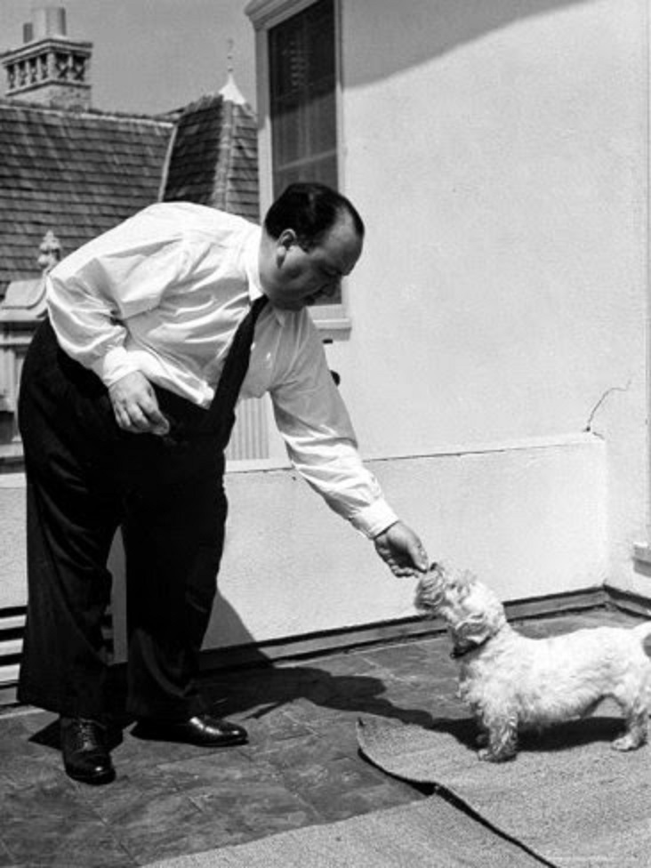Director Alfred Hitchcock standing outside at home feeding his sealyham terrier Mr. Jenkins.