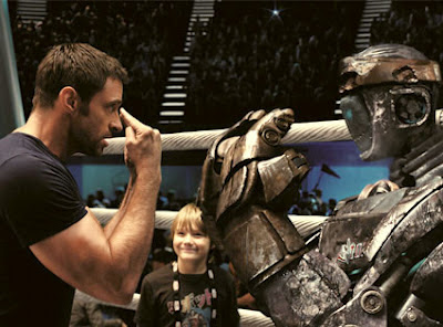 Hugh Jackman in a boxing ring with his robot boxer and he is point with both hands to his eyes and the robot is mimicking him.