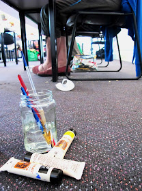 Jar with paint brushes, and paint tubes on a floor under a table at a workshop.