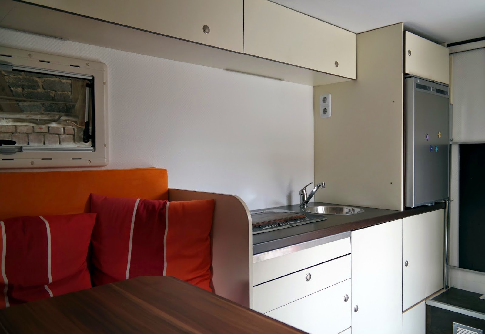 Küchenzeile über den Esstisch fotografiert. Am Ende der Zeile der 110 Liter Kühlschrank mit kleinem Gefrierfach. Darüber und darunter Platz für Lebensmittel. Ganz am Boden befinden sich die beiden Varta Batterien und nebenan in dem braunen "Tritt" Spannungswandler, Sicherungen und Controller für die Solarpanelen.