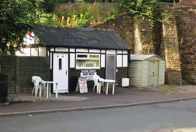 Chip shop hut with white tables and chairs outside in the shadow of railway bridge.